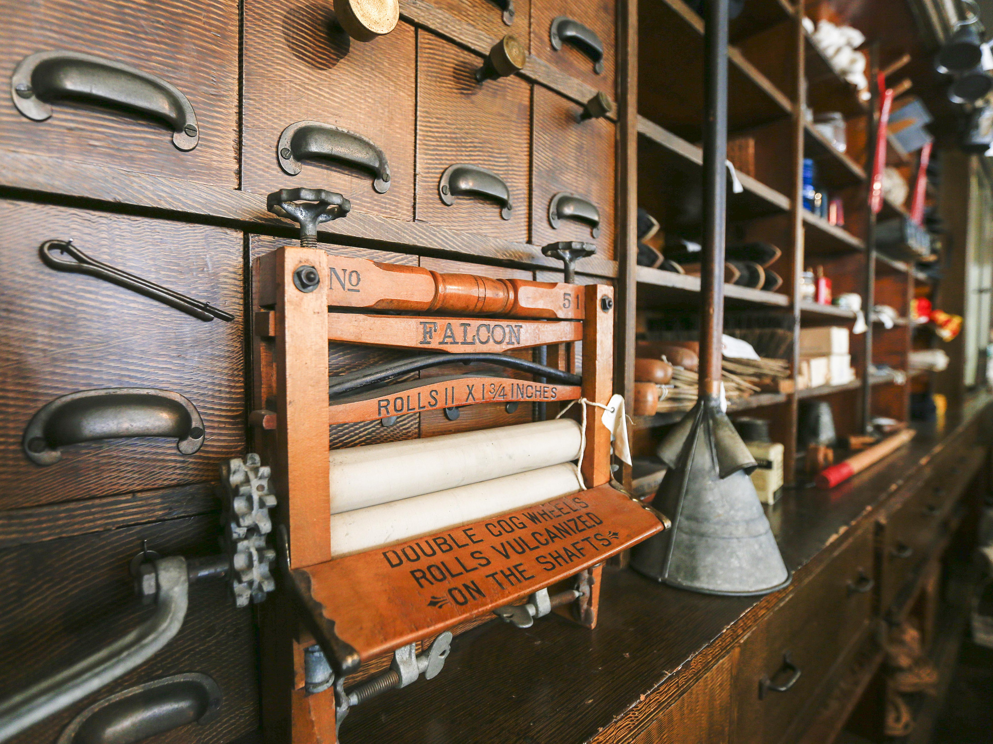 Close-up of a vintage store counter featuring a wooden drawer unit with metal handles, a "Falcon" brand washing machine wringer with orange text reading "ROLLS 11 X 2 INCHES" and "DOUBLE COG WHEELS ROLLS VULCANIZED ON THE SHAFTS", and various metal tools and parts.