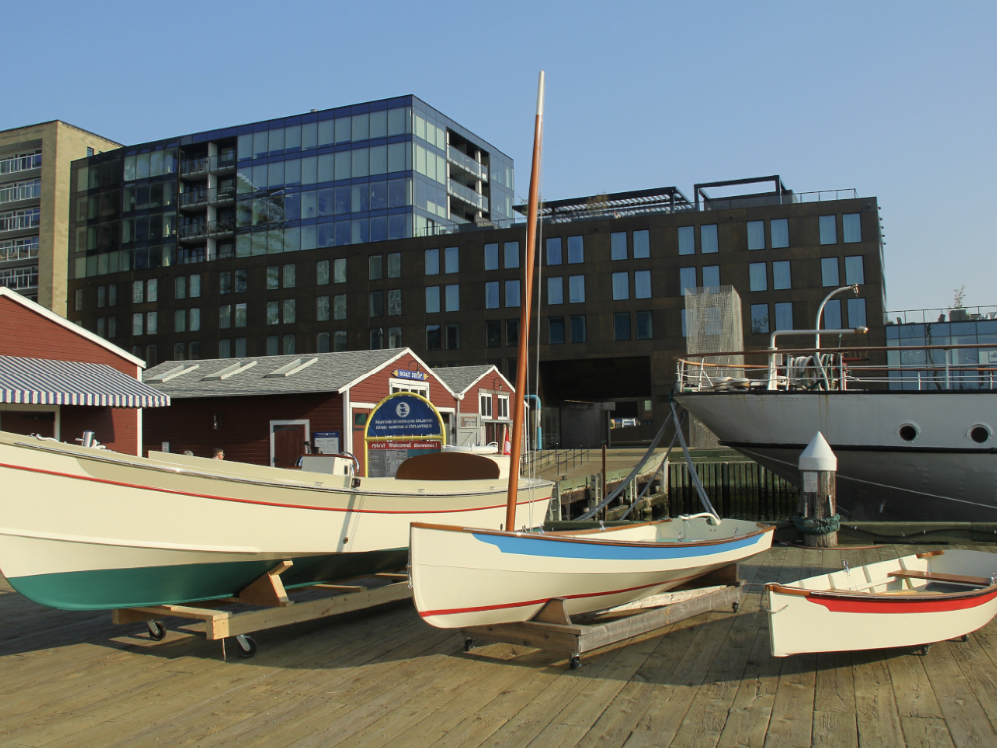 Three wooden boats of different sizes are displayed on wooden cradles on a boardwalk. The largest boat has a cream hull with a teal bottom, the middle one has a cream hull with a blue stripe, and the smallest is cream with a red stripe. Behind them are several small, red-roofed buildings and a large, modern building with dark glass windows. Part of a larger, white ship is visible on the right.
