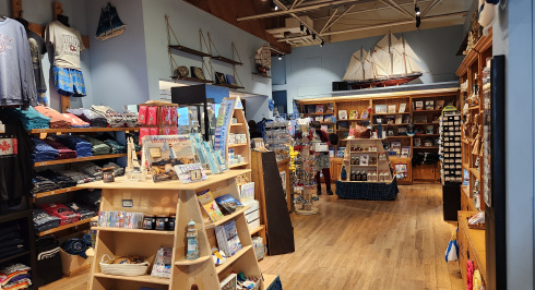  A spacious gift shop with a high ceiling and wooden floor, filled with maritime-themed merchandise. In the foreground, there are wooden display shelves with various items like books, small decorative lighthouses, and baskets. To the left, racks of clothing are visible, featuring t-shirts and shorts. In the background, more shelves line the walls, displaying books and other souvenirs.