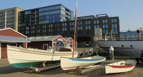 Three wooden boats of different sizes are displayed on wooden cradles on a boardwalk. The largest boat has a cream hull with a teal bottom, the middle one has a cream hull with a blue stripe, and the smallest is cream with a red stripe. Behind them are several small, red-roofed buildings and a large, modern building with dark glass windows. Part of a larger, white ship is visible on the right.