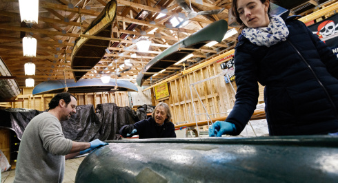 An active, low-angle shot inside a rustic wooden boatbuilding workshop shows three museum staff members or conservators wearing blue protective gloves as they carefully restore or examine the hull of an overturned green canoe. Hanging directly overhead from the exposed wooden ceiling trusses are several vintage canoes, including a classic birchbark or canvas-covered model and a dark green canoe. The workshop is brightly lit by overhead industrial lights, with exposed wooden stud walls.