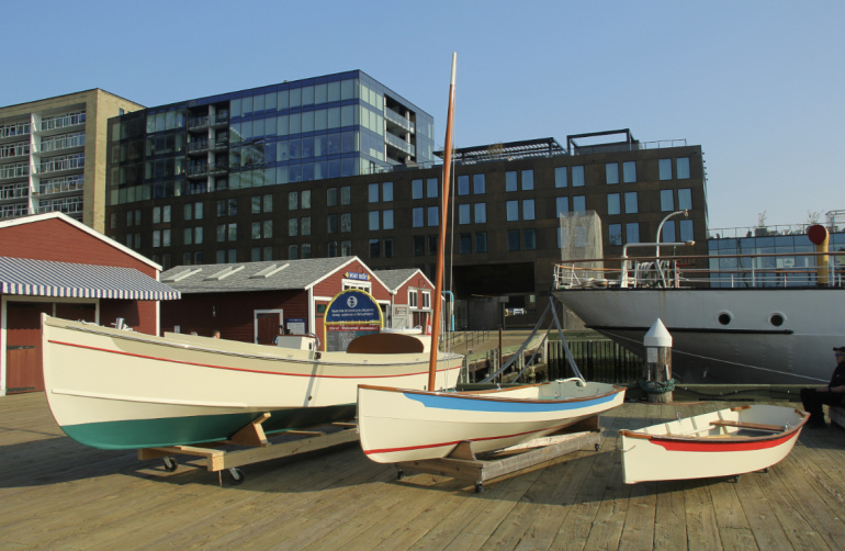 Three wooden boats of different sizes are displayed on wooden cradles on a boardwalk. The largest boat has a cream hull with a teal bottom, the middle one has a cream hull with a blue stripe, and the smallest is cream with a red stripe. Behind them are several small, red-roofed buildings and a large, modern building with dark glass windows. Part of a larger, white ship is visible on the right.