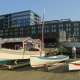 Three wooden boats of different sizes are displayed on wooden cradles on a boardwalk. The largest boat has a cream hull with a teal bottom, the middle one has a cream hull with a blue stripe, and the smallest is cream with a red stripe. Behind them are several small, red-roofed buildings and a large, modern building with dark glass windows. Part of a larger, white ship is visible on the right.
