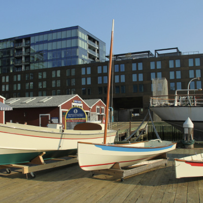 Three wooden boats of different sizes are displayed on wooden cradles on a boardwalk. The largest boat has a cream hull with a teal bottom, the middle one has a cream hull with a blue stripe, and the smallest is cream with a red stripe. Behind them are several small, red-roofed buildings and a large, modern building with dark glass windows. Part of a larger, white ship is visible on the right.
