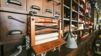 A close-up view of a vintage washing machine wringer, labeled 'FALCON,' with wooden rollers and metal components. The wringer is positioned in front of a multi-drawer wooden cabinet with small, dark metal handles. The drawers and other shelves in the background are filled with various unidentified objects.
