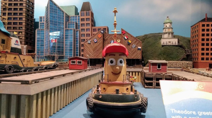  A large model of Theodore Tugboat, the main character from the Canadian children's television show, is displayed in a miniature version of a city harbor. Theodore is a smiling tugboat with a red cap. The background features models of various buildings, including a prominent hill with a white clock tower (resembling the Halifax Citadel Clock Tower), suggesting the setting is based on Halifax, Nova Scotia.