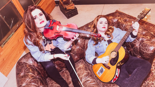 Cassie & Maggie laying on a leather sofa holding their instruments. 